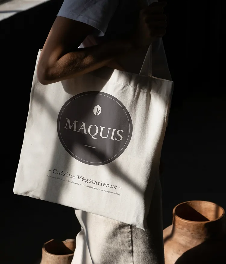 Eine Stofftasche mit weißem Maquis-Logo auf schwarzem Grund, darunter der Schriftzug ‚Cuisine Végétarienne‘ und die Adresse des Restaurants in Hamburg.