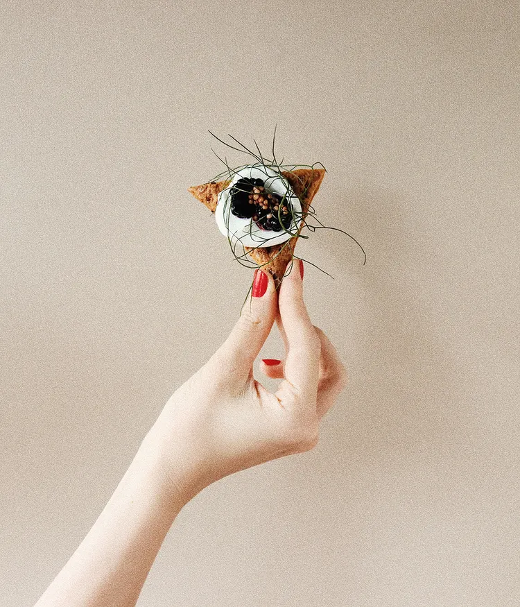 Eine Hand vor beigem Hintergrund hält einen exquisit angerichteten Cracker mit Crème fraîche und feiner Garnitur.