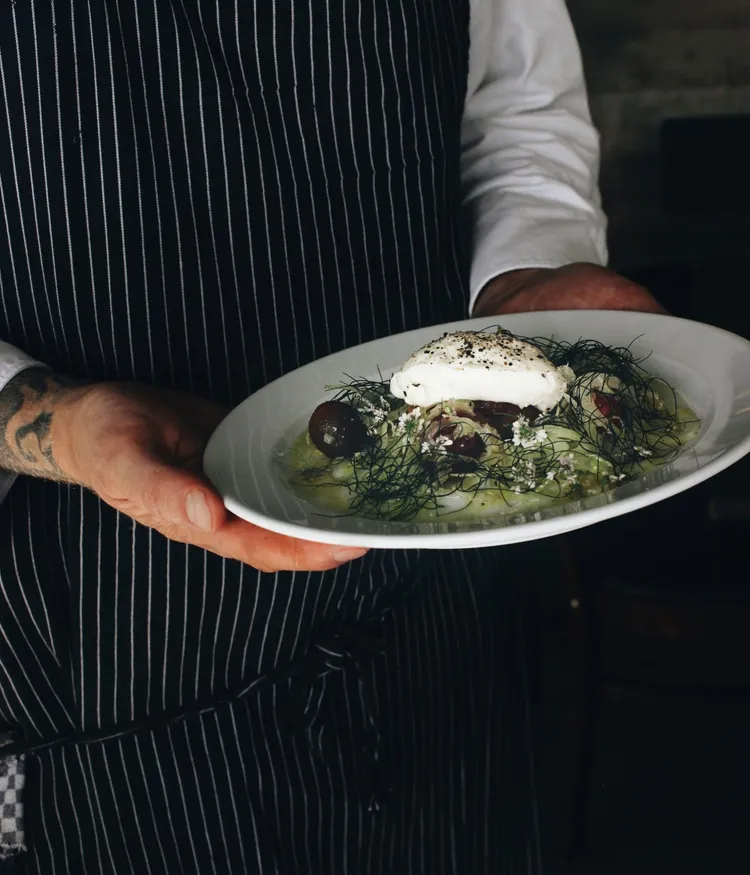 Die Hände eines Koches mit gestreifter Schürze präsentieren ein schmackhaft aussehendes Gericht aus der Küche-Maquis.