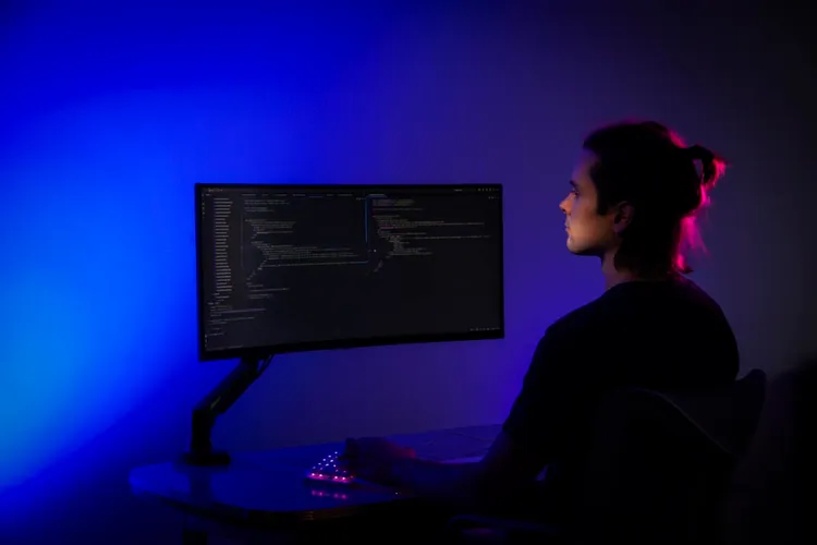 Eine Person mit einem Dutt sitzt vor einem Monitor in einem dunkel beleuchteten Raum. Der Monitor zeigt Code an. Das Licht ist in Blau- und Violetttönen gehalten, was eine futuristische Atmosphäre schafft.