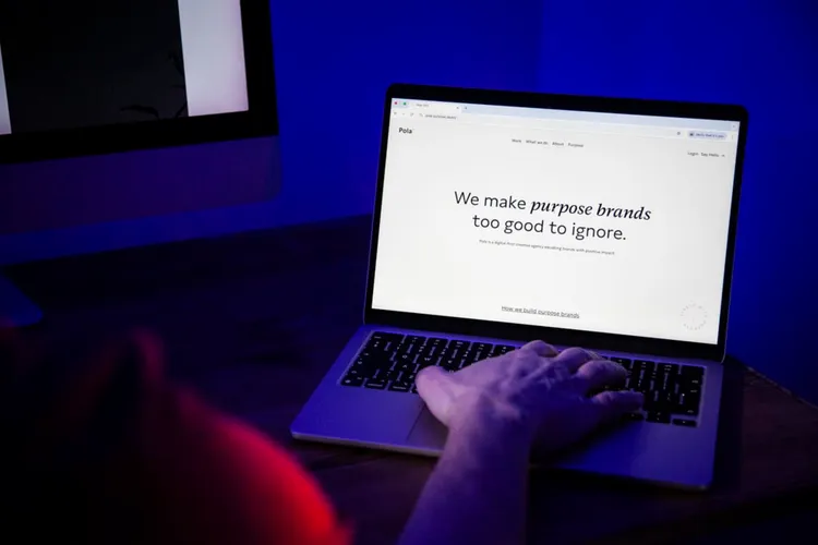 Eine Person sitzt vor einem Laptop in einem dunklen Raum mit blauem Licht. Auf dem Bildschirm ist eine Webseite geöffnet, die den Text 'We make purpose brands too good to ignore.' zeigt.