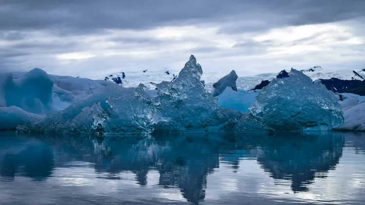 Schwimmende Eisformationen auf ruhigem Wasser, im Hintergrund schneebedeckte Berge.